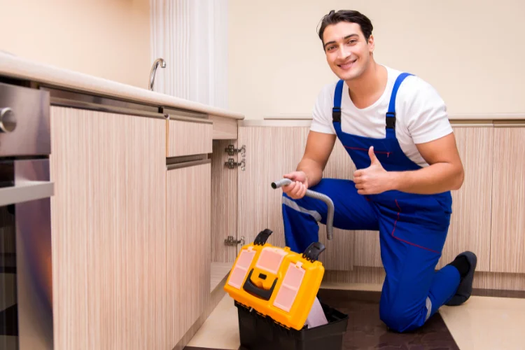 Smiling plumber giving thumbs up while repairing kitchen plumbing with a toolbox